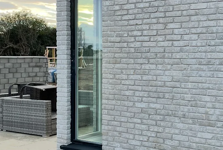 Limewashed white brick elevation with a tall slim window and paved terrace at sunset