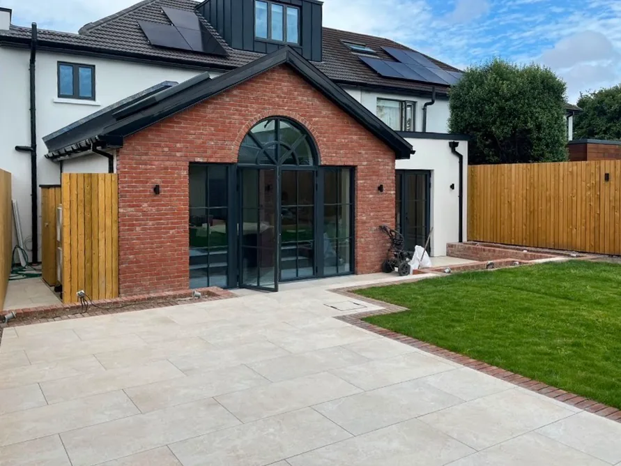 Red brick gable with an arched steel-framed door set, white rendered house behind and travertine paving